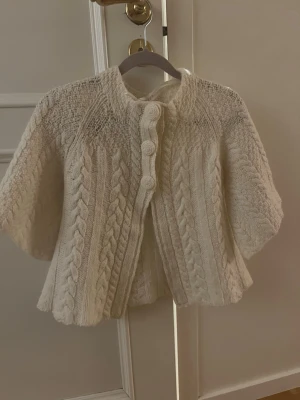 Vit trekvarts stickad kofta med flätmönster - Superfin vit stickad kofta med korta ärmar och stora stickade knappar framtill. Koftan har ett klassiskt flätmönster och är gjord i ett mjukt, tjockt garn som känns riktigt cozy. Perfekt att slänga över en t-shirt eller klänning för extra värme.