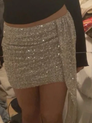 Glittrig silver kortkjol med slits - Säljer en glittrig silverfärgad kortkjol med snygg slits och draperad detalj framtill. Kjolen har en tight passform och är perfekt för dig som vill sticka ut. Ingen synlig logga eller märke.