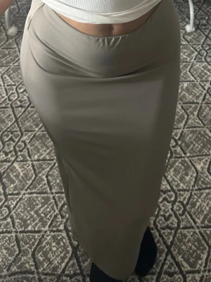 Beige långkjol i stretchigt material - Säljer en mörk beige långkjol med figurnära passform. Kjolen är enkel och stilren utan mönster eller detaljer, tillverkad i ett stretchigt syntetmaterial som sitter snyggt på kroppen. Perfekt att matcha med crop tops eller stickade tröjor.