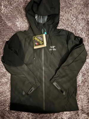 Svart Arc'teryx vindjacka Gore-Tex - Snygg svart vindjacka från Arc'teryx med huva och vattentätt Gore-Tex-material. Jackan har hel dragkedja framtill, justerbara muddar och diskret logga på bröstet. Perfekt för dig som vill ha en stilren och funktionell jacka.