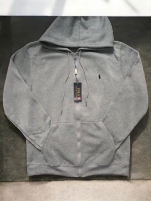  Polo Ralph Lauren - Snygg grå hoodie från Polo Ralph Lauren med dragkedja framtill, huva med dragsko och klassisk broderad logga på bröstet. Mjuk bomullsblandning och känguruficka gör den både bekväm och stilren. Perfekt för en avslappnad streetwear-look.