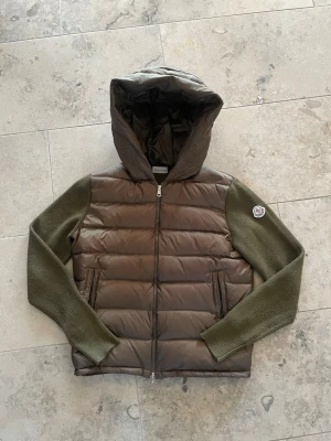 Moncler brun/grön dunjacka med huva - Snygg brun dunjacka från Moncler med stickade olivgröna ärmar och stor huva. Jackan har dragkedja framtill, två fickor och Moncler-logga på ärmen. Perfekt för kalla vinterdagar och riktigt trendig look.