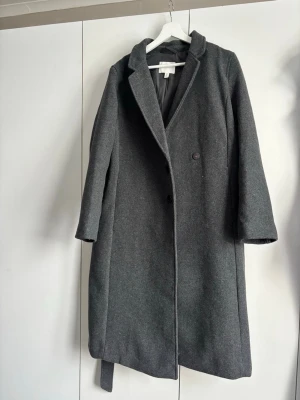 Mörkgrå ullkappa med bälte - Stilren mörkgrå ullkappa från H&M med klassisk krage och två knappar framtill. Kappan har ett matchande bälte i midjan och raka ärmar. Den är använd endast en gång, så skick som ny. Storlek 42 men passar snarare en 36/38 (liten i storleken)