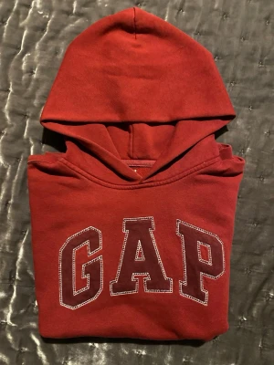 Röd hoodie från GAP med logga - Säljer en klassisk röd hoodie från GAP med stor vit och svart broderad logga på bröstet. Hoodien har huva och en stor magficka framtill. Tillverkad i mjukt bomullsmaterial, perfekt för en avslappnad och snygg stil.