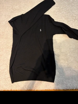 Svart stickad tröja från Ralph Lauren - Klassisk svart stickad tröja från Ralph Lauren med rund halsringning och diskret broderad logga på bröstet. Tröjan har långa ärmar och ribbade muddar vid hals, ärmslut och nederkant. Perfekt basplagg för en stilren look.