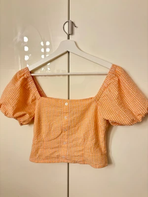 Korall rutig topp - Superfin croppad topp från EDITED i korall- och vitrutigt mönster. Toppen har puffiga korta ärmar, knappar framtill och en fyrkantig urringning. I strl 36, passar mig me xs-m