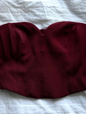 Vinröd korsett-topp med dragkedja - Snygg vinröd korsett-topp med hjärtformad urringning och markerade sömmar. Toppen är axelbandslös och har dragkedja bak för enkel påklädning. Perfekt att styla med höga jeans eller kjol för en trendig look.