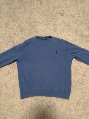 Blå tröja från Polo Ralph Lauren - Klassisk blå tröja från Polo Ralph Lauren med rund halsringning och diskret broderad logga på bröstet. Tröjan har ribbade muddar vid ärmslut och nederkant. Perfekt för en stilren och enkel look. Den är i bra skick utan deffekter! Tveka inte på att höra av er!
