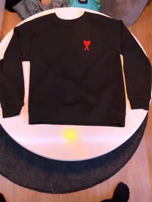 Svart sweatshirt från Ami Paris - Svart sweatshirt från Ami Paris med röd broderad logga i form av ett hjärta och bokstaven A på bröstet. Klassisk rund halsringning och ribbade muddar vid ärmslut och nederkant. Perfekt för en stilren och trendig look.