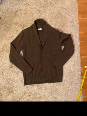 Brun Lindbergh kofta - Snygg brun kofta med sjalkrage och fem stora knappar framtill. Långärmad modell i mjukt material, perfekt för lager på lager. Enkel och stilren design som passar till det mesta.