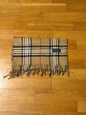 halsduk Burberrys - Klassisk beige halsduk från Burberrys i 100% lammull med ikoniskt rutmönster i svart, vitt och rött. Halsduken har fransar i ändarna och är tillverkad i England. Perfekt accessoar för att lyfta din outfit.