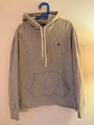 Grå hoodie från Polo Ralph Lauren - Snygg grå hoodie från Polo Ralph Lauren med klassisk huva och vita dragsnören. Har en känguruficka framtill och den ikoniska lilla loggan broderad på bröstet. Perfekt för en chill och stilren look.
