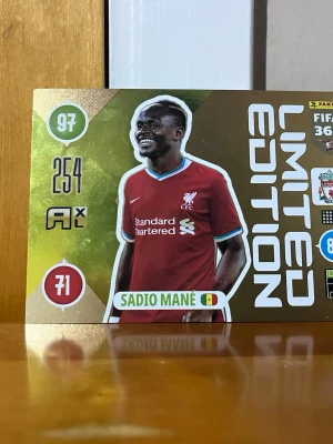 Limited Edition Sadio Mané kort - Limited Edition samlarkort från Panini med Sadio Mané i Liverpool-tröja. Kortet har guldfärgad bakgrund, vita och gröna detaljer samt spelarens namn och statistik. Perfekt för fotbollssamlare och fans av Liverpool.