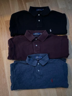 Pikétröjor från Polo Ralph Lauren - Säljer tre klassiska pikétröjor från Polo Ralph Lauren i svart, vinröd och mörkblå. Alla har krage, knappar framtill och den ikoniska broderade loggan på bröstet. Perfekta för en clean och stilren look.