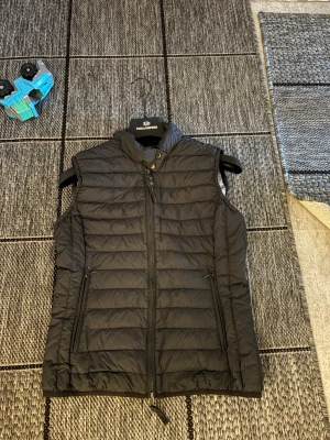 Svart dunväst från Parajumpers - Snygg svart dunväst från Parajumpers med hög krage och dragkedja framtill. Västen har två praktiska fickor med dragkedja och ett broderat Parajumpers-märke i nacken. Perfekt för lager-på-lager och riktigt stilren.