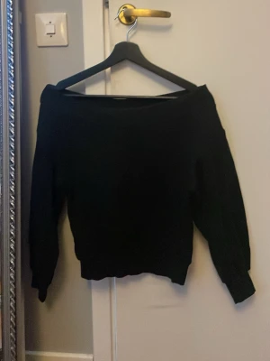 Svart off shoulder mjukis tröja  - Snygg svart mjukis tröja med off shoulder-modell och långa ärmar. Tröjan har ribbade muddar vid ärmslut och nederkant, vilket ger en skön och trendig passform. Perfekt för dig som gillar en enkel men stilren look.