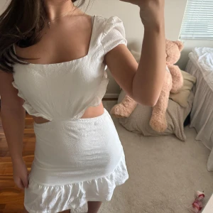 Vit kort klänning med öppen rygg - Supersöt vit kort klänning med puffärmar, volangkant nertill och öppen rygg med knytband. Klänningen har en fyrkantig ringning och är figurnära i modellen. Perfekt för studenten men även som en sommar klänning.