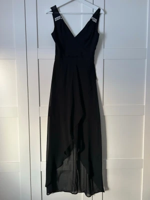 Svart långklänning med pärldetaljer - Elegant svart långklänning med v-ringning och pärldetaljer på axlarna. Klänningen har ett lager av tunn, flytande polyester och asymmetrisk nederkant som ger en snygg siluett. Perfekt för dig som vill sticka ut med stilrena detaljer. OBS! Fattas 2 stenar på ena sidan (syns på bild 4) annars i mycket bra skick. 