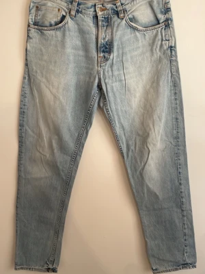 Ljusblå jeans från Nudie Jeans - Säljer ett par ljusblå jeans från Nudie Jeans med klassisk femficksmodell och raka ben. Jeansen har snygga slitningar och kontrastsömmar samt Nudie-logga på bakfickan. Tillverkade i bomull för en skön och avslappnad look.