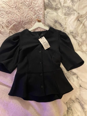 Svart puffärmad blus från Zara - Stilren svart blus från Zara med puffiga korta ärmar och markerad midja. Blusen har rund halsringning och knäppning framtill med svarta knappar. Materialet är lätt strukturerat och ger en elegant look. Perfekt för att lyfta vilken outfit som helst.