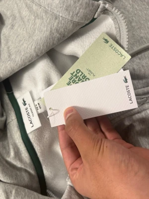 Grå zip-hoodie från Lacoste - Snygg grå hoodie från Lacoste med dragkedja och klassisk krokodillogga på bröstet. Hoodien har huva med dragsko och är tillverkad i mjukt material som känns skönt mot huden. Perfekt för en chill och stilren look.