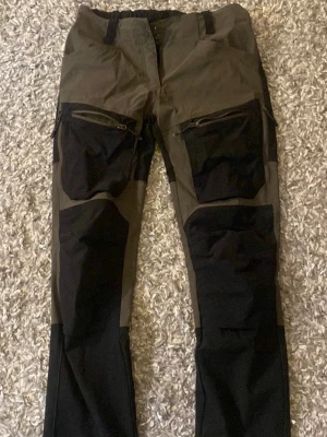 Gröna och svarta cargobyxor med dragkedjor, Whistler - Snygga cargobyxor i grönt och svart med flera praktiska fickor och dragkedjor på benen. Byxorna har förstärkta partier och är tillverkade i slitstarkt material, perfekta för en aktiv livsstil. Rak passform och bälteshällor upptill.