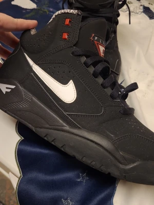 Svarta Nike Air Flight sneakers - Säljer ett par svarta Nike Air Flight sneakers med vit swoosh och röda detaljer. Skorna har högt skaft, svart sula och mönstrat innerfoder i vitt och svart. Klassisk streetstil med perforerade partier och robust design.