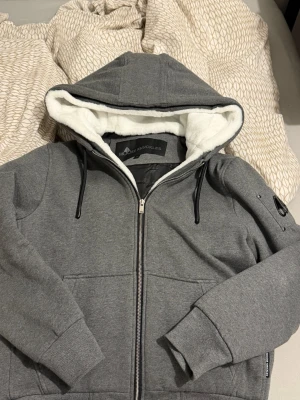 Moose Knuckles jacka  - Snygg grå jacka från Moose Knuckles med dragkedja, stor huva fodrad i vit fuskpäls och svarta dragsnören. Hoodien har ribbade muddar, fickor framtill och en detalj på ärmen. Perfekt för dig som vill ha både stil och komfort.