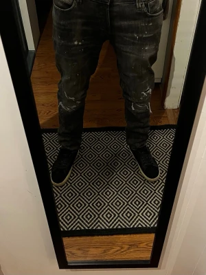 Svarta distressed jeans med stänk - Säljer ett par svarta jeans med coola slitningar och vita färgstänk. Jeansen har en rak passform och klassiska fem fickor. Perfekta för dig som gillar en edgy streetstil. Materialet är denim. Jack and Jones. Aldrig använda. 