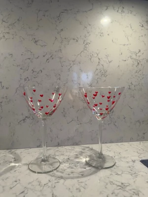 Två cocktailglas med röda hjärtan - Säljer två genomskinliga cocktailglas med röda hjärtan som dekoration. Glasen har klassisk form med bred kupa och lång smal fot. Perfekta för drinkar eller som söt inredningsdetalj.