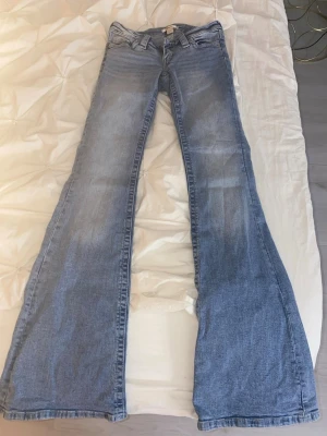 Blå bootcut jeans med broderade fickor - Säljer ett par ljusblå bootcut jeans med snyggt slitna detaljer och broderade bakfickor. Jeansen har låg midja och klassisk femficksdesign. Perfekta för dig som gillar retrostil och vill ha ett par jeans med extra vidd nedtill.