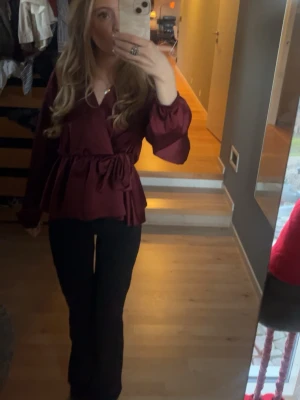 Vinröd omlottblus med volang - Säljer en vinröd omlottblus med volangdetaljer och knytband i midjan. Blusen har långa, puffiga ärmar och v-ringning. Perfekt för dig som gillar trendiga och feminina plagg. Storlek XS/S. Satin material