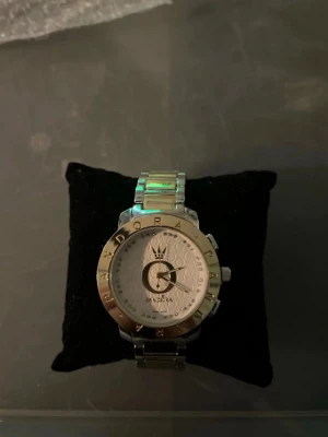 Silverfärgad klocka från Pandora - Stilren klocka från Pandora i silverfärg med metallarmband och rund urtavla. Urtavlan har Pandoras logga och detaljer i guldton samt markerade siffror runt kanten. Länken är robust och har en graverad Pandora-logga på spännet.