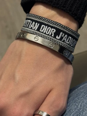 Christian Dior J'Adior armband - Snyggt armband från Christian Dior med texten 'J'Adior' i vitt på svart tygband med dekorativa pärldetaljer och tofsar. Perfekt för att ge din outfit en exklusiv touch. Kombineras snyggt med andra smycken. Snabb leverans. Skriv gärna om du har frågor.