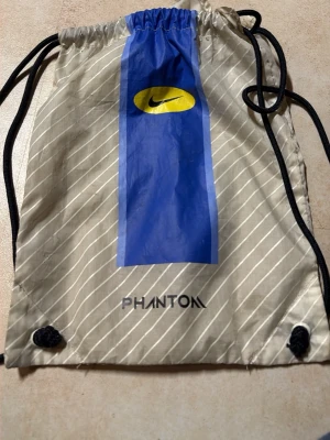 Nike Phantom fotbollsskor beige/blå - Säljer ett par Nike Phantom fotbollsskor i beige med blå och svarta detaljer samt gul Nike-logga. Skorna har snörning och är tillverkade i syntetmaterial med textilinslag. Medföljer originalpåse med Phantom-tryck och Nike-logga.