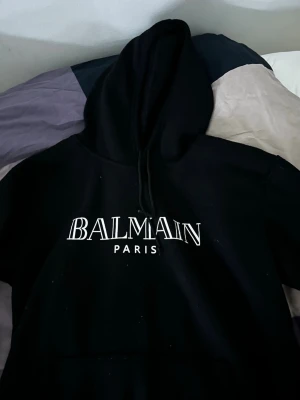 Svart Balmain Paris hoodie - Snygg svart hoodie från Balmain Paris med stor vit logga på bröstet. Klassisk huva med dragsko och känguruficka framtill.  Mjuk och skön bomullskänsla.