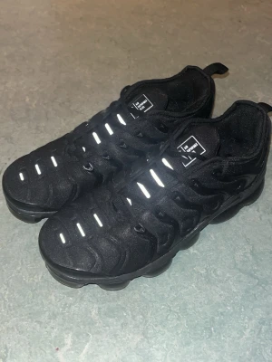 Nike Air VaporMax Plus svarta sneakers - Nike Air VaporMax Plus sneakers i helsvart med reflekterande detaljer på ovandelen. Skorna har en unik, vågig design och en robust sula med synliga luftbubblor för extra komfort. Materialet är en mix av textil och syntet. Perfekta för dig som gillar en sportig och modern look