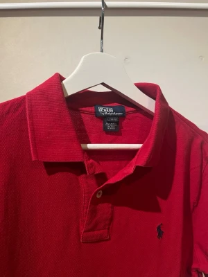 Pikétröja från Polo Ralph Lauren - Klassisk röd pikétröja från Polo Ralph Lauren med broderad logga på bröstet. Tröjan har krage, korta ärmar och knappar vid halsen. Perfekt för en stilren och sportig look. (Storlek L i barn så passar XS!)