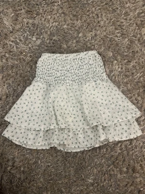 Vit blommig volangkjol - Supersöt kort kjol i vitt med små blommor och volang. Kjolen har smock i midjan för skön passform och dubbla lager tyg som ger en luftig känsla. Perfekt för varma dagar och enkel att matcha med olika toppar.