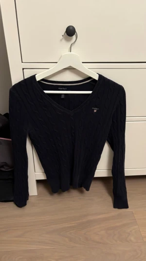 Mörkblå kabelstickad tröja Gant - Snygg mörkblå kabelstickad tröja från Gant med V-ringning och diskret logga broderad på bröstet. Tröjan har lång ärm och ribbade muddar vid ärmslut och nederkant. Perfekt för en klassisk och stilren look.
