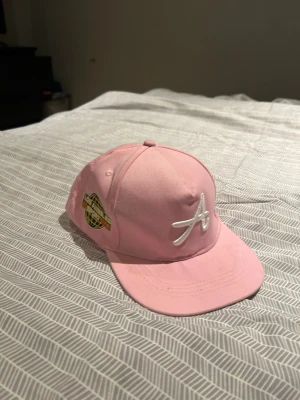 Rosa keps med vit broderad A-logga - Säljer en snygg rosa keps med vit broderad A-logga framtill och en guldfärgad etikett på sidan. Klassisk form med böjd skärm och justerbar baksida. Perfekt accessoar för en trendig streetwear-look.
