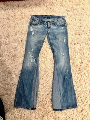 Blå bootcut jeans med slitningar - Ett par ljusblå bootcut jeans med slitna detaljer och råa kanter. Jeansen har klassisk femficksdesign, låg midja och utställda ben. Snygga kontrastsömmar och patch baktill i midjan. Perfekta för en avslappnad och trendig look.