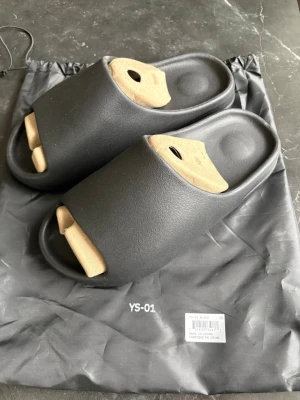 Svarta Yeezy ”slides” YS-01  - Säljer ett par svarta slides med chunky, räfflad sula och öppen tå. Tofflorna har en minimalistisk design och är tillverkade i ett mjukt, skumliknande material för extra komfort. Perfekta att glida i hemma eller till och från gymmet. Kom med bud.