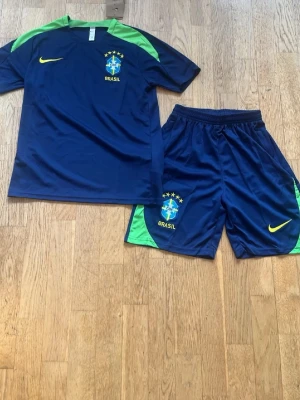 Blått Brasilien fotbollskit Nike Rabbat om man köper direkt - Snyggt blått Brasilien fotbollskit från Nike med matchande tröja och shorts. Tröjan har gröna axelpartier, gul Nike-logga och Brasilien-emblem på bröstet. Shortsen har gröna detaljer på sidorna och samma emblem. Perfekt för fotbollsträning eller match.