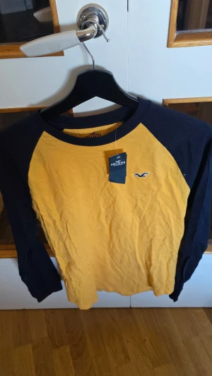 Gul och marinblå långärmad Hollister - Snygg långärmad tröja från Hollister i gul med marinblå ärmar och krage. Klassisk raglanmodell med broderad logga på bröstet. Perfekt för en avslappnad och trendig stil.