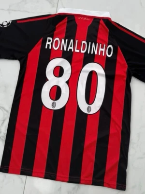 Fotbollströja - Säljer en klassisk AC Milan matchtröja med Ronaldinho och nummer 80 på ryggen. Tröjan är röd och svart med vita detaljer, Adidas logga och Bwin sponsortryck. Tillverkad i lätt och ventilerande ClimaCool-material, perfekt för fotbollsträning eller samling.
