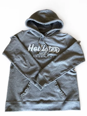 Grå hoodie från Hollister - Snygg grå hoodie från Hollister med broderad logga och text på bröstet. Klassisk känguruficka framtill och justerbar huva med dragsko. Mjuk insida och ribbade muddar vid ärmar och nederkant. Perfekt för en avslappnad och trendig look. Pris kan diskuteras, hör av dig om du har några frågor!