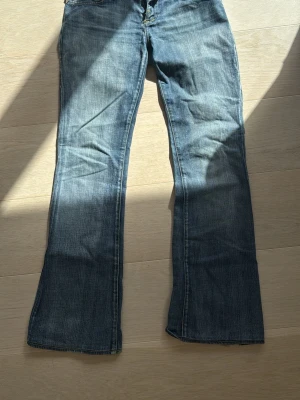Blå bootcut jeans med fickdetaljer - Snygga blå bootcut jeans med klassisk femficksdesign och gul söm på bakfickorna. Jeansen har låg midja och lätt slitning för en avslappnad look. 