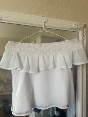 Vit offshoulder volangtopp - Söt vit offshoulder topp med volang och smock upptill. Tunt, luftigt material och axelband. Perfekt för varma dagar och ger en gullig look. Toppen har två väldigt små fläckar, som knappt syns. 