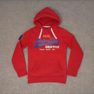 Röd Superdry hoodie - Snygg röd hoodie från Superdry med blått och vitt tryck på bröstet.  Hör av vid frågor!